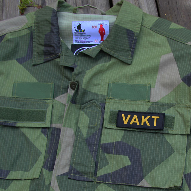 Ett VAKT Kardborremärke monterat på locket på en bröstficka på en M90 kamouflage jacka.