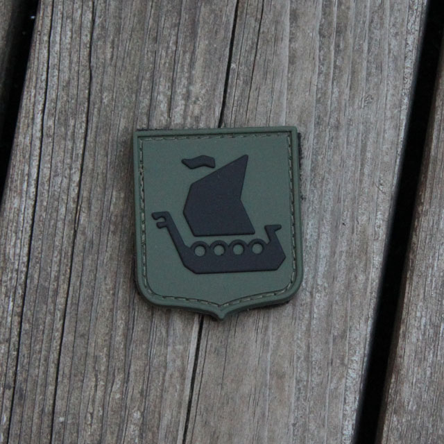 Ett Vikingship Shield Hook PVC Green/Black Patch mot en träbakgrund.