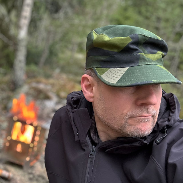 Patrol Cap M90 from TAC-UP GEAR on a model in the Swedish forest seen at a slight angle with a fire in the background