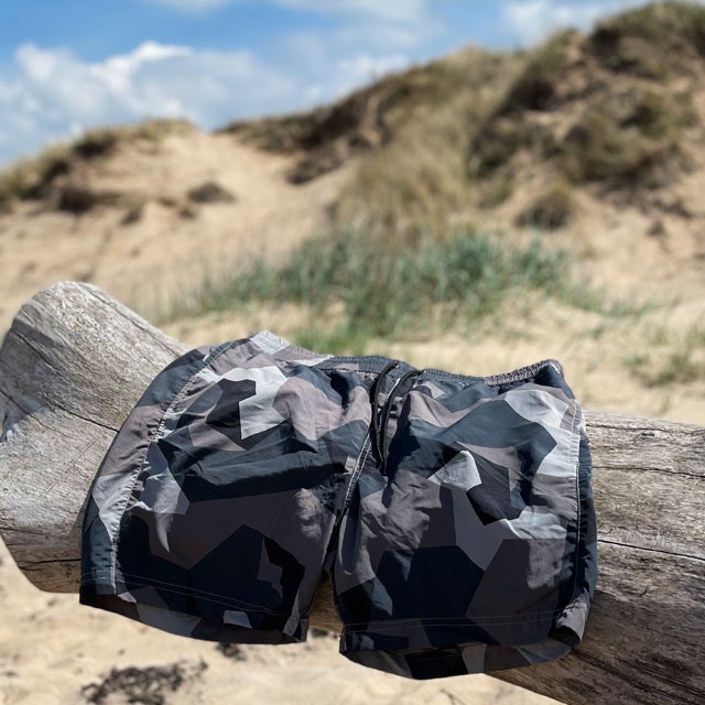 A pair of POSEIDON Swim Shorts M90 Grey from TAC-UP GEAR hanging on a log on a beach