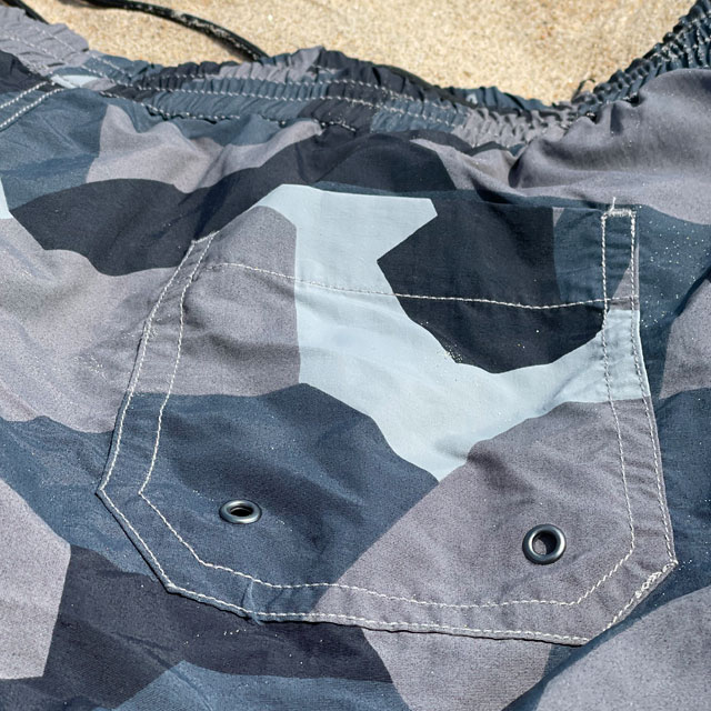 The backpocket on a pair of POSEIDON Swim Shorts M90 Grey lying flat on the beach