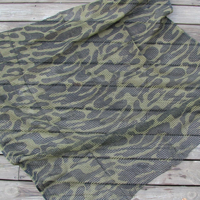 A Scrim Scarf Black/OD Camo laying against a wooden bakground showing its pattern.