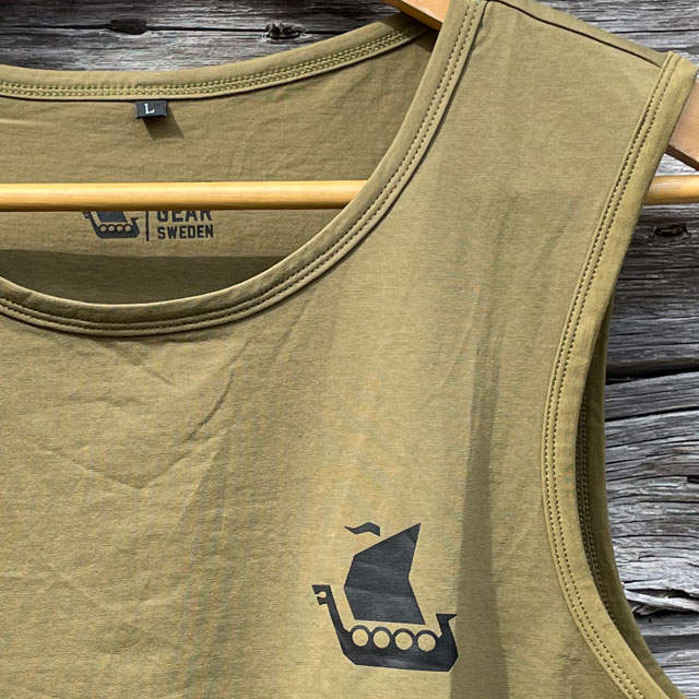 A Tank Top Khaki Green from TAC-UP GEAR on a hanger with old woodhouse background with focus on the neck and breast print