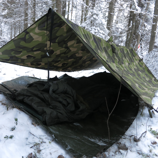 Winter usage of the Tarp Poncho M90 seen here as roof over sleepingbag and sleepingmat on the snow covered ground.