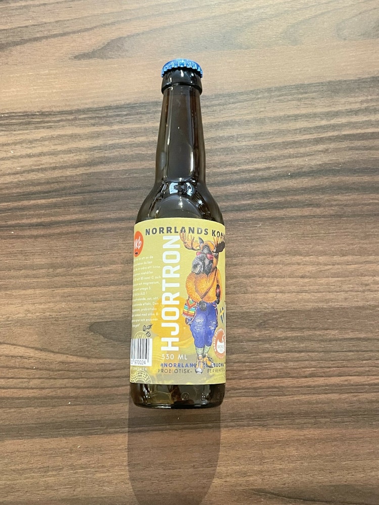 kombucha i brun glasflaska med gulgrön ettikett med en älg på. Smak av hjortron