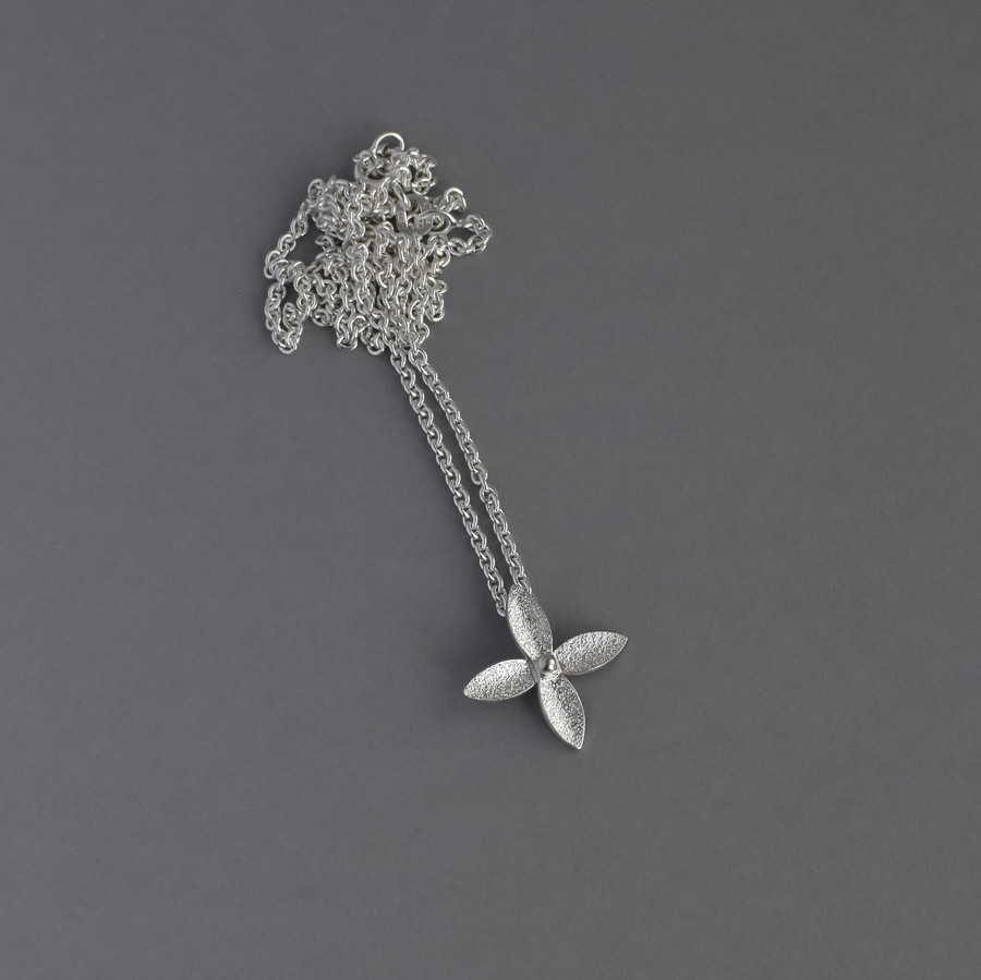 Hänge av silver (925) med en 45 cm lång silverkedja. Hänget består av en liten blomma med fyra spetsiga kronblad och en kula i mitten. Blomman är ca 18 cm i diameter. Hängets yta har en lite skrovlig 